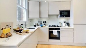 A modern kitchen with white cabinets, a gas stove, a microwave, and a sink. There is a breakfast setup with croissants and orange juice on the counter. - PBC | Luxury One Bedroom Apartment in Central London, Fitzrovia (GT12) (London)