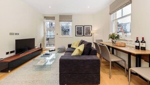 A modern living room with a flat-screen TV, a sofa, a coffee table, and a dining area with a table and chairs. - PBC | Luxury One Bedroom Apartment in Central London, Fitzrovia (GT12) (London)