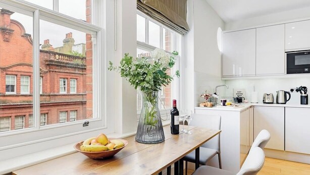 A modern kitchen with white cabinets, a wooden dining table, and a vase with flowers. - PBC | Luxury One Bedroom Apartment in Soho GT13 (London)
