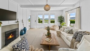 A spacious living room with a fireplace, a large sofa, a wooden coffee table, and a view of the outdoors through large windows. - Drift @ 5 Mile Bay (Taupo)