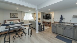 A modern living room with a sofa, a dining table with stools, a kitchen with a sink and cabinets, and a TV mounted on the wall.