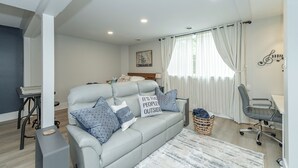 A modern living room with a grey sofa, a small table, and a chair. There is a window with curtains and a painting on the wall.