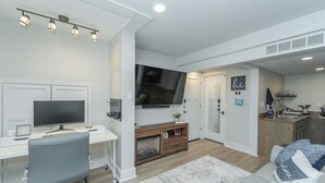 A modern living room with a white desk, grey office chair, and a large flat-screen TV.