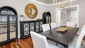 Un comedor con mesa de madera oscura, sillas blancas, candelabro y elementos decorativos, incluyendo un espejo en forma de sol y armarios negros.