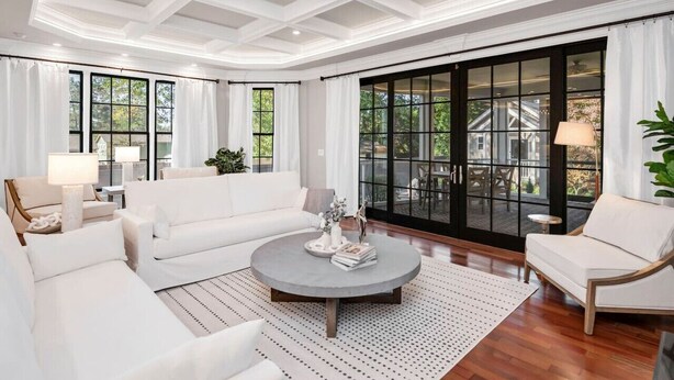Un salón amplio con un sofá blanco, una mesita redonda y ventanas grandes que ofrecen vistas a una casa y follaje verde.