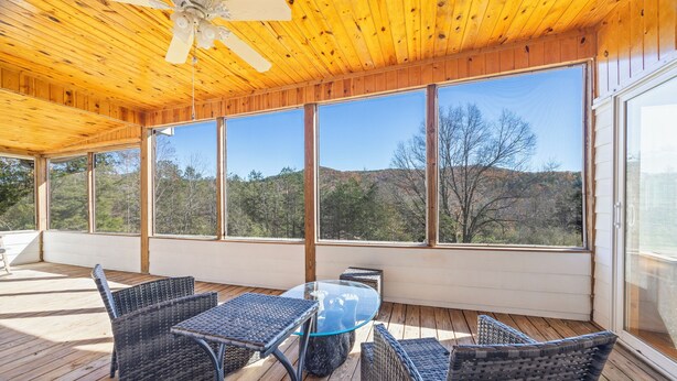 Un espacio al aire libre cubierto con mobiliario de mimbre, una mesa de vidrio y ventanas amplias con vistas a árboles y colinas.