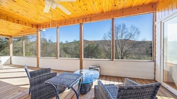 Un espace extérieur abrité, aménagé avec des meubles en osier, une table en verre et de grandes fenêtres offrant une vue sur les arbres et les collines.