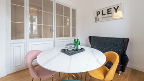 A dining area with a white marble table, pink and yellow chairs, and a black velvet chair with pink piping. A sign reading "PLEY BOUTIQUE" is on the wall.