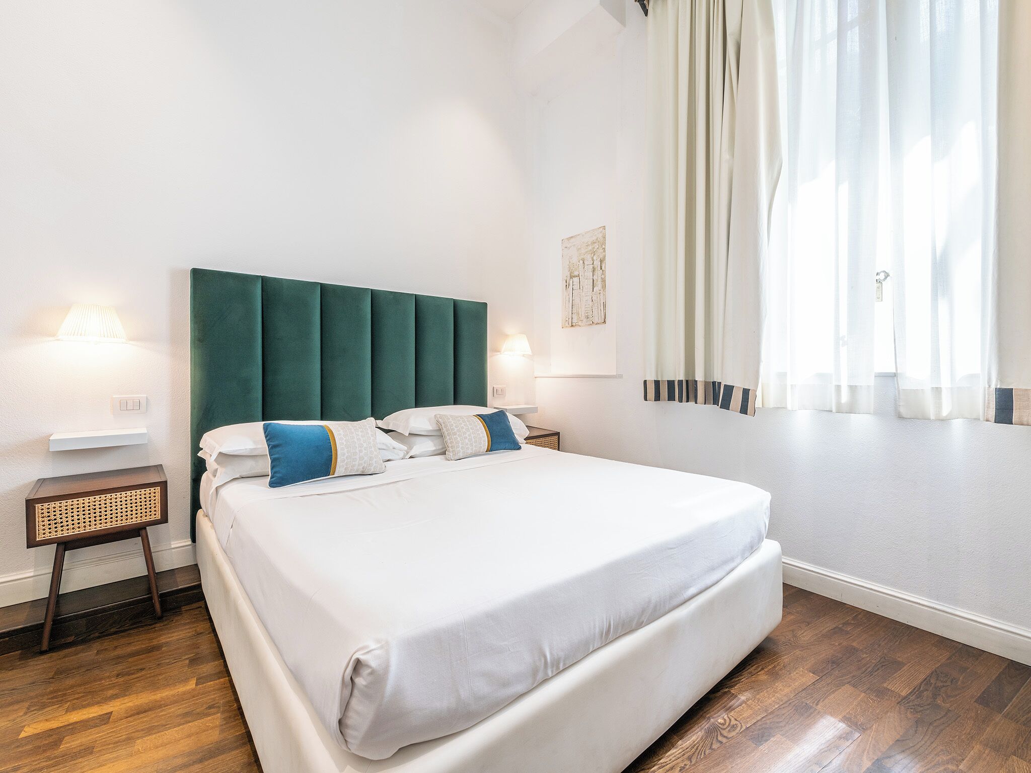 Una cama bien hecha con sábanas blancas y almohadas azules, un cabecero tapizado en verde, una mesita de noche con una lámpara, y una ventana con cortinas traslúcidas.