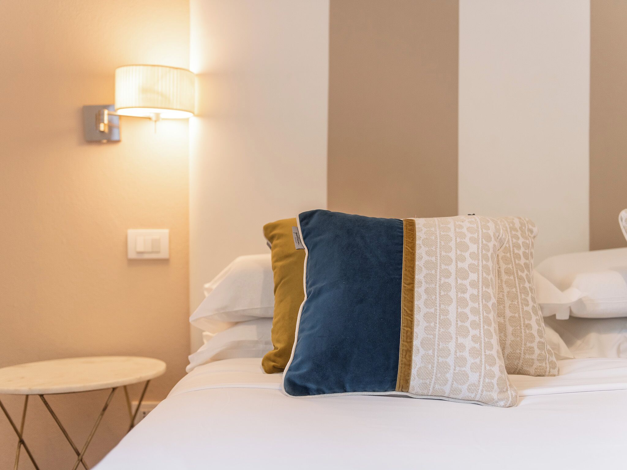 Una habitación de hotel con cama, almohadas, una mesita redonda y una lámpara de pared.