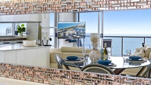 Un espace repas moderne offrant une vue sur le paysage urbain, doté d’un grand téléviseur à écran plat et d’un balcon avec une balustrade.