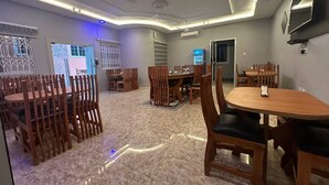 A dining area with wooden chairs and tables, a ceiling fan, and a TV mounted on the wall. - Senalli Hotel (Koforidua)