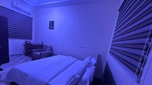 A bedroom with a bed, a chair, a desk, and a window with blinds. - Senalli Hotel (Koforidua)