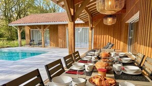 A dining table set with plates, bowls, and croissants, with a pool and wooden building in the background.