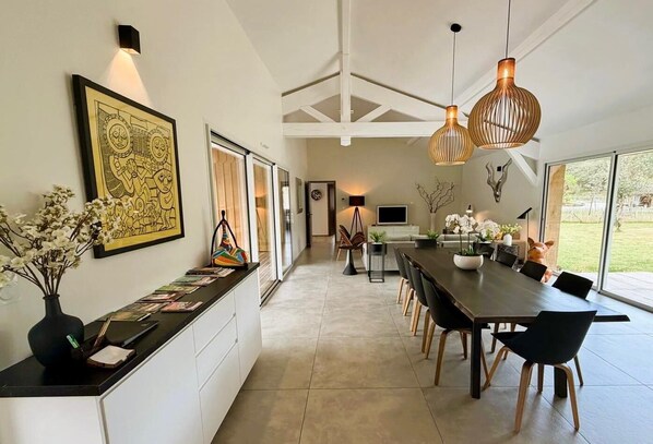 A modern kitchen with a long countertop, dining area with chairs, pendant lights, and a large window.
