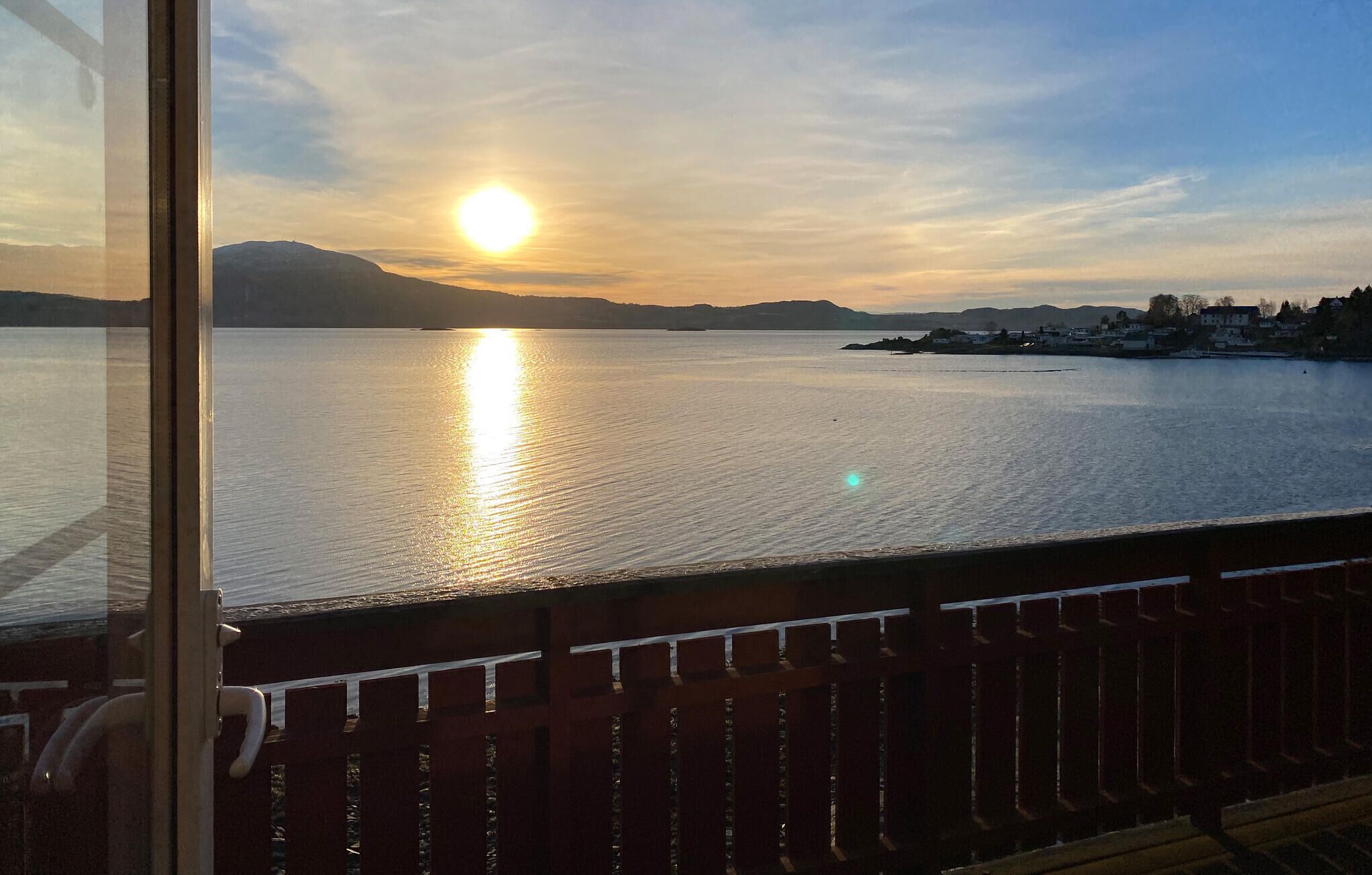 Une vue d’un lac paisible au coucher du soleil, avec une chaîne de montagnes lointaine et une petite ville sur la rive opposée.