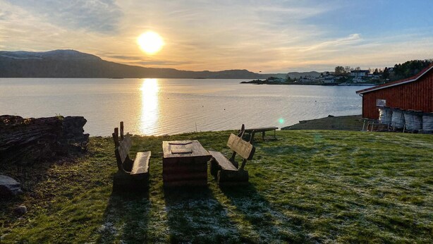 A lakeside view with a wooden bench and picnic table, a red building, and mountains in the background. - Beach front home in Frosta with WiFi (Frosta)
