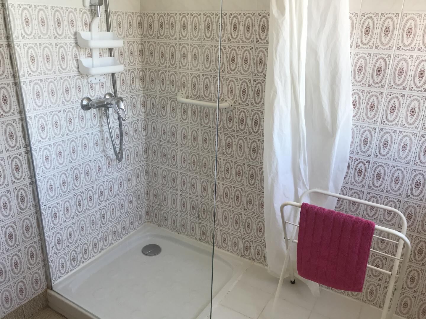 A shower area with a patterned tile wall, a white showerhead, and a pink towel on a metal rack.
