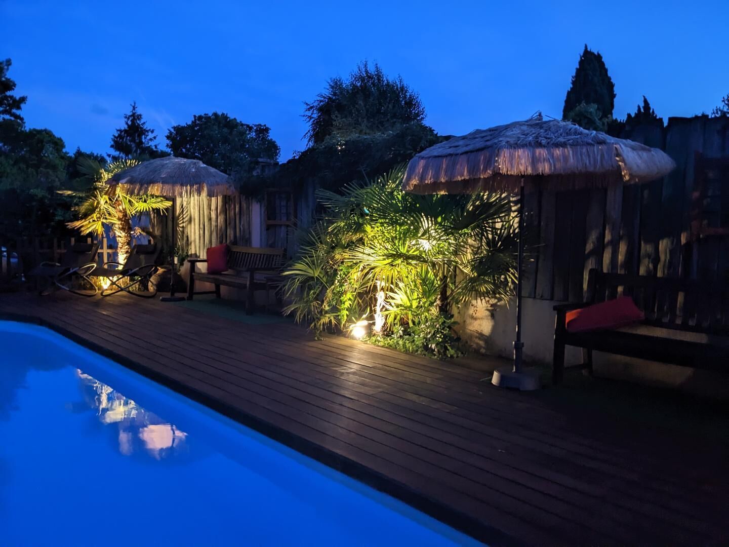 A poolside area with wooden decking, lounge chairs, and thatched umbrellas.