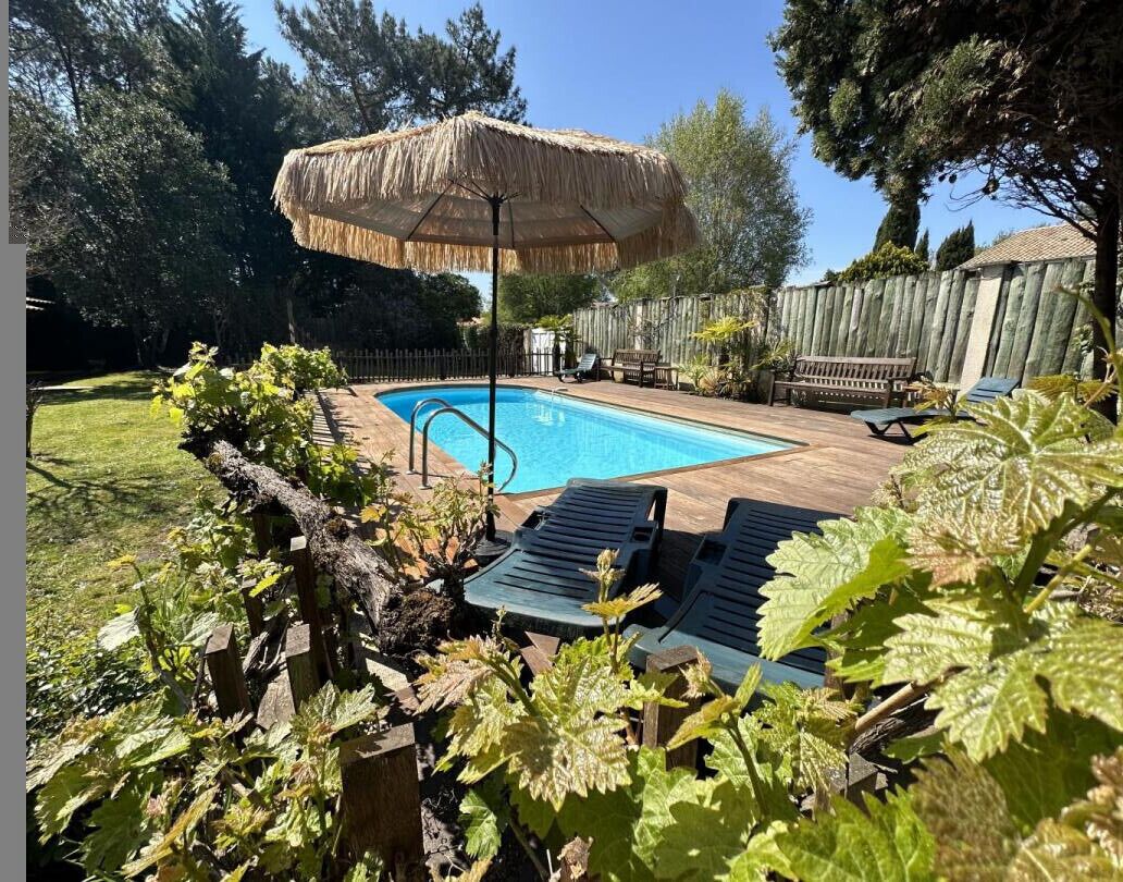 Um quintal com piscina, cadeiras de descanso, um guarda-sol de palha e uma cerca de madeira.
