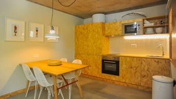 Een kleine keuken met houten kastjes, een magnetron en een spoelbak. Er staat een eettafel met stoelen, en aan de muur hangen prenten.
