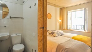 Une salle de bain équipée de toilettes, d’un lavabo et d’une porte en bois donnant sur une chambre avec un lit, une fenêtre et une lampe fixée au mur.