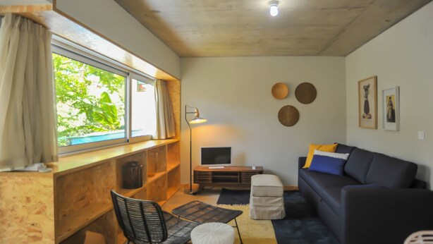 Un salon accueillant avec un bureau en bois, une chaise noire, un canapé bleu foncé agrémenté de coussins, un petit pouf blanc et un téléviseur à écran plat posé sur un meuble en bois.
