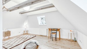 A spacious attic room with wooden beams, a small table, a chair, a radiator, and a window.