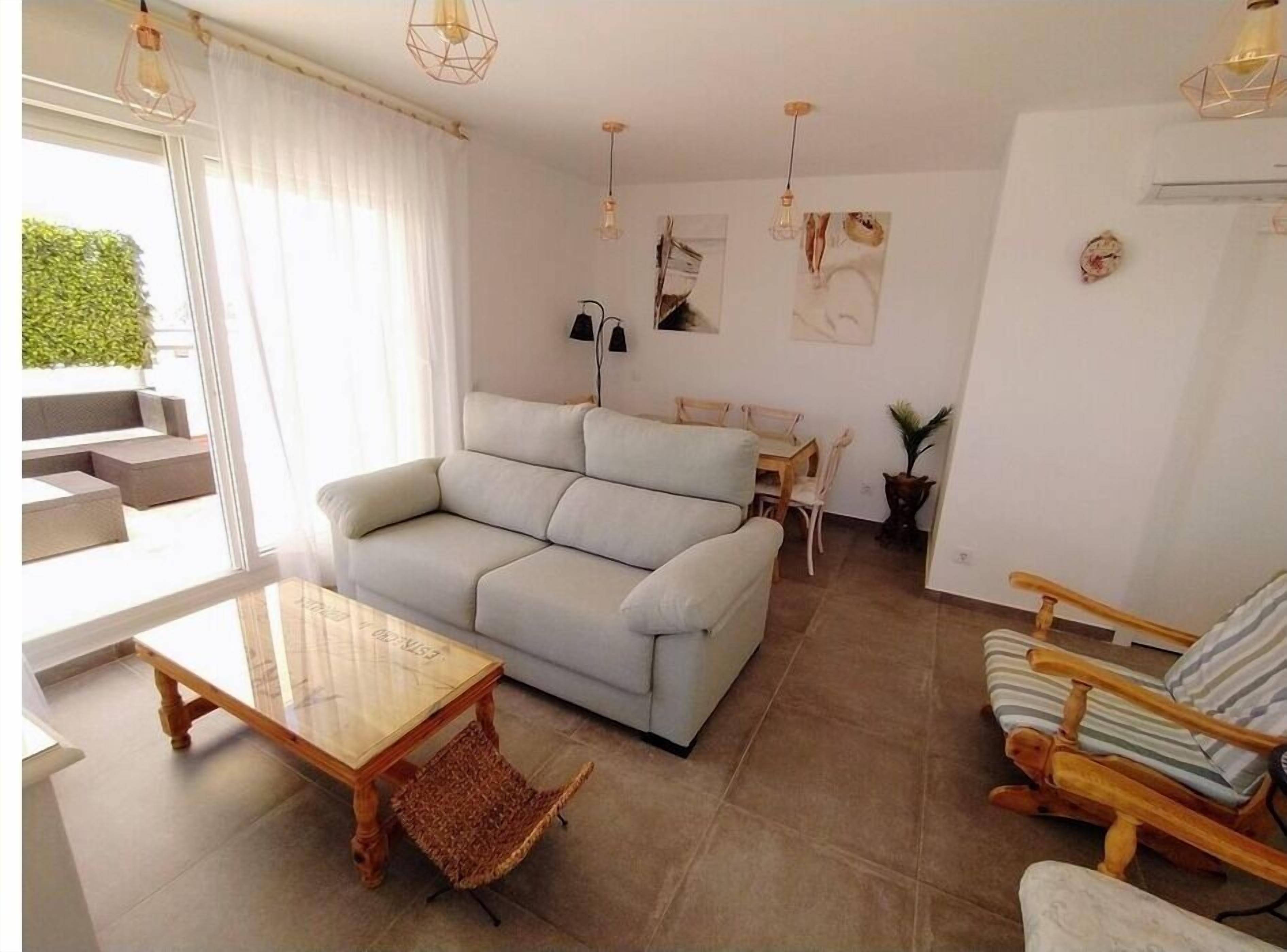 Een moderne woonkamer met een grijze bank, een houten salontafel, een gestreepte fauteuil en een eethoek met een kleine tafel en stoelen.