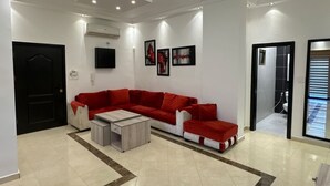 A modern living room with a red sofa, a coffee table, and a TV mounted on the wall. - Central Secured Compound w/ Hill-view Terrace (Amman)