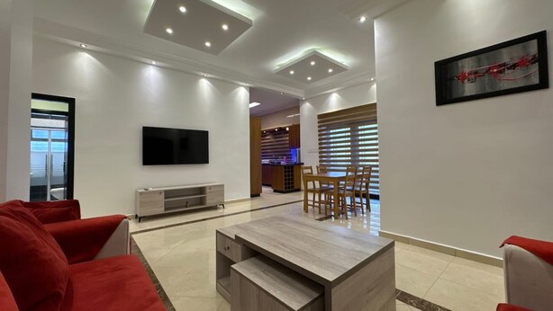 A modern living room with a TV, a wooden coffee table, red sofas, and a dining area with a table and chairs. - Central Secured Compound w/ Hill-view Terrace (Amman)