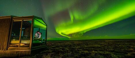 A glass-roofed structure with a person inside, set against a backdrop of the Northern Lights.