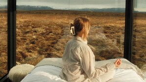 A person sitting on a bed in a room with large windows, overlooking a barren landscape. - Tiny Glass Lodge (Selfoss)