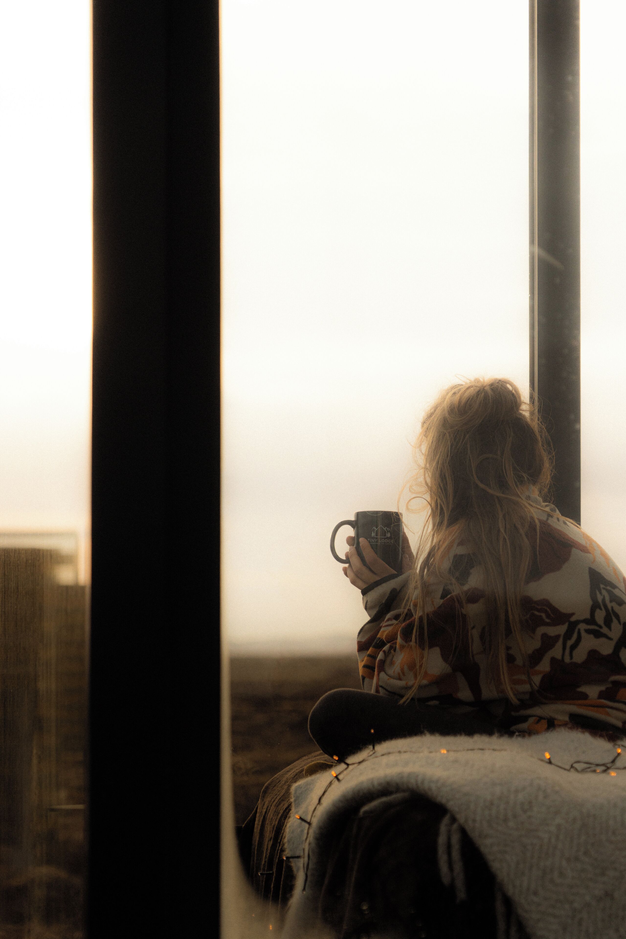 A person sitting by a window with a cup, wearing a patterned top, and a blanket.