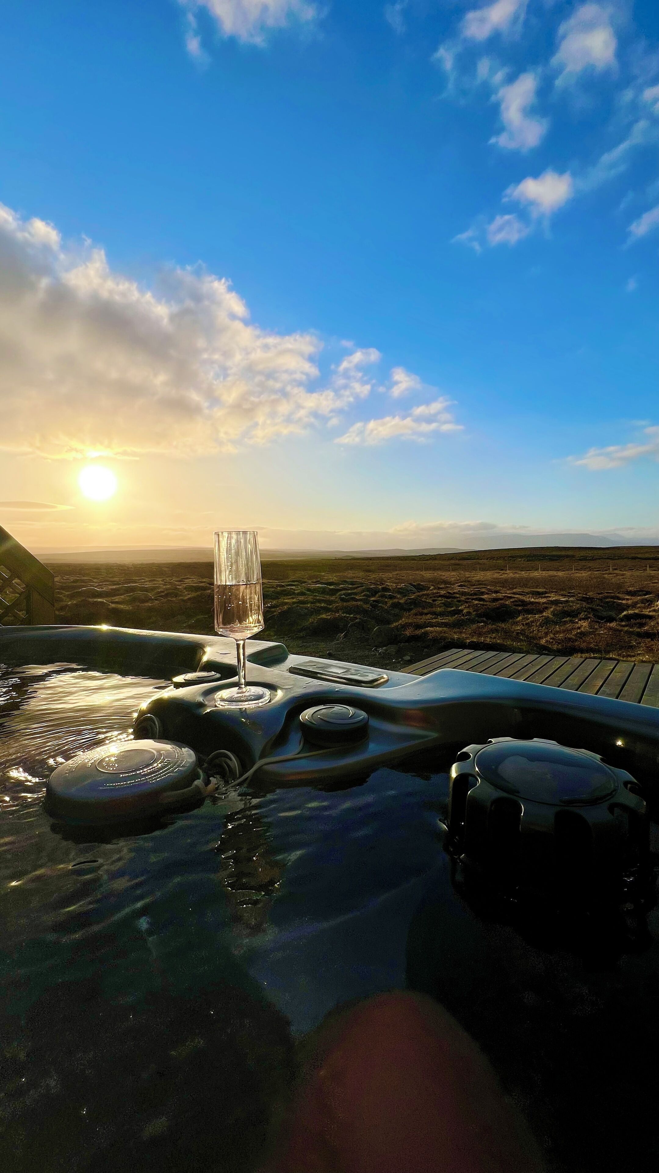 A hot tub with a glass of water and a remote control, surrounded by a wooden deck, with a view of the landscape and the sun setting in the background.