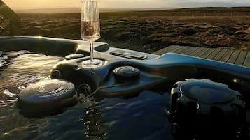 A hot tub with a glass of water and a remote control, surrounded by a wooden deck, with a view of the landscape and the sun setting in the background.