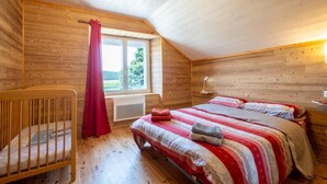 A cozy wooden room with a sloped ceiling, a window with a view, a bed with a striped blanket, a crib, and a bedside lamp.