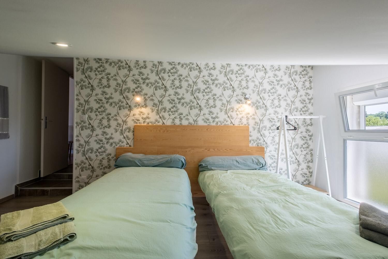 Une chambre avec deux lits, un papier peint à motifs floraux, une tête de lit en bois et une fenêtre donnant sur un beau paysage.