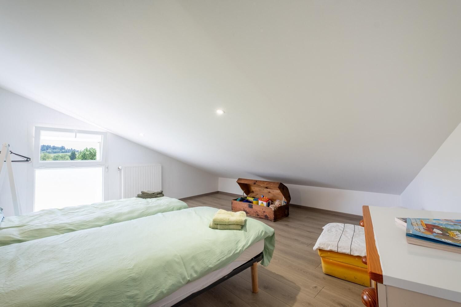A bedroom with two beds, a wooden chest of drawers, a window with a view, and a table with a book.