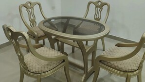 A dining area with a round glass table and four chairs, a flat-screen TV mounted on the wall, and a tiled floor.