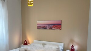 A neatly made bed with pillows and a blanket, a bedside table with a red decorative item, a painting of a sunset on the wall, and a hanging light fixture.