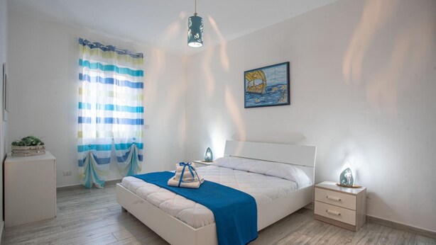 A bedroom with a bed, bedside tables, a hanging light fixture, a window with curtains, and a painting on the wall.