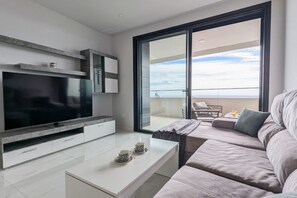 A modern living room with a flat-screen TV, a sofa, a coffee table, and a balcony with a view.