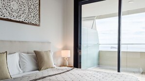 Un dormitorio con una cama grande, una mesita de noche con lámpara, un adorno de pared y vista al cielo a través de una puerta de cristal.