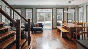 Amplia sala de estar con una escalera de madera, un conjunto de sofás, una mesa de comedor con sillas y ventanas grandes que ofrecen vistas al exterior.