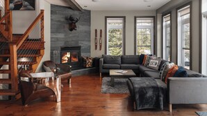 Una sala de estar moderna con chimenea de piedra, una escalera de madera y ventanas amplias que ofrecen vistas al exterior.