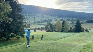 Camp de golf amb un jugador preparant-se per fer una rematada, envoltat d'arbres i un paisatge ondulat al fons.
