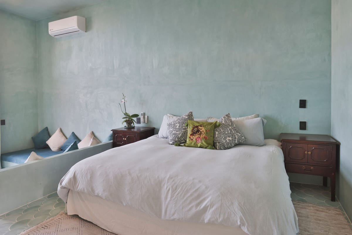 Un dormitorio con cama, mesitas de noche, aire acondicionado instalado en la pared y una ventana pequeña con vistas.