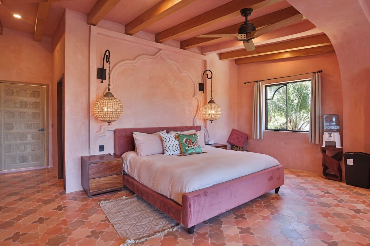 Un dormitorio con una cama rosa, alfombra estampada, mesitas de noche de madera y una ventana con cortinas.