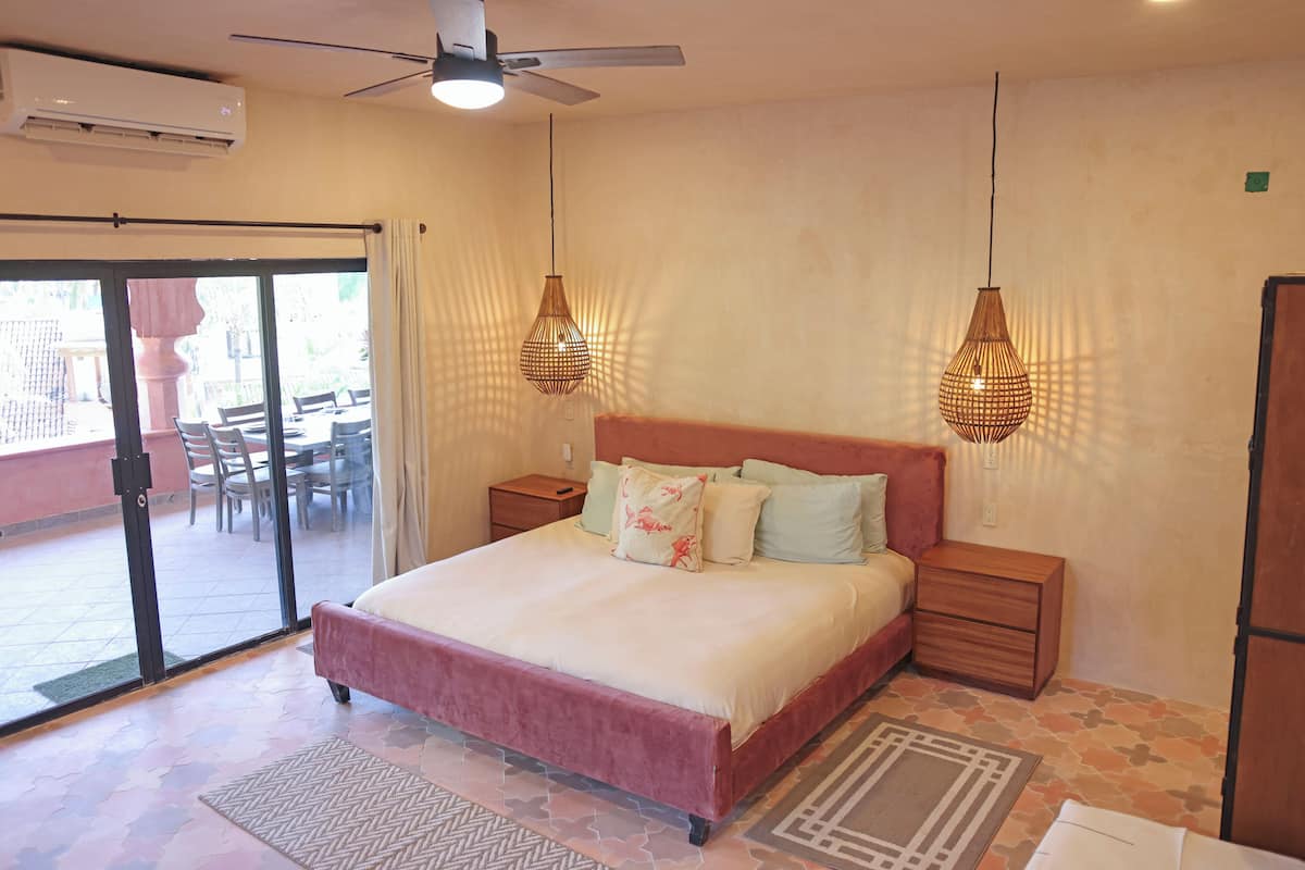 Un dormitorio con cama, mesitas de noche, ventilador de techo y vistas a un patio exterior a través de un ventanal.